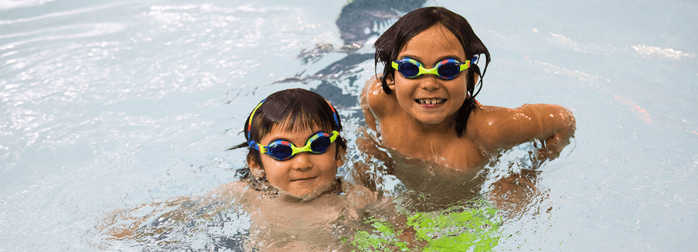 Two children in the pool with swim goggles on smiling.