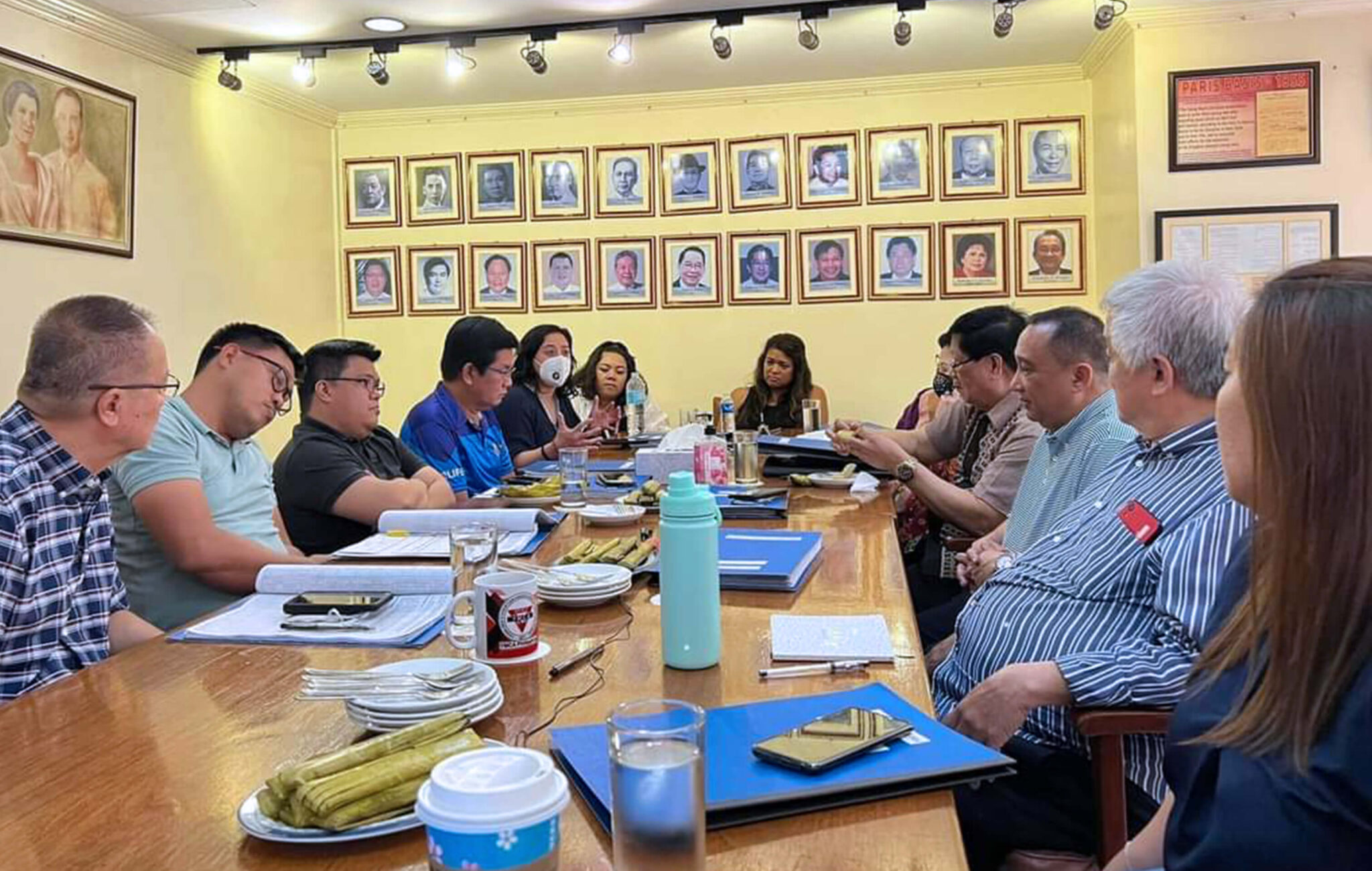 A group of people are sitting at a long table during a board meeting. There are two rows of pictures hung on the yellow wall behind them.