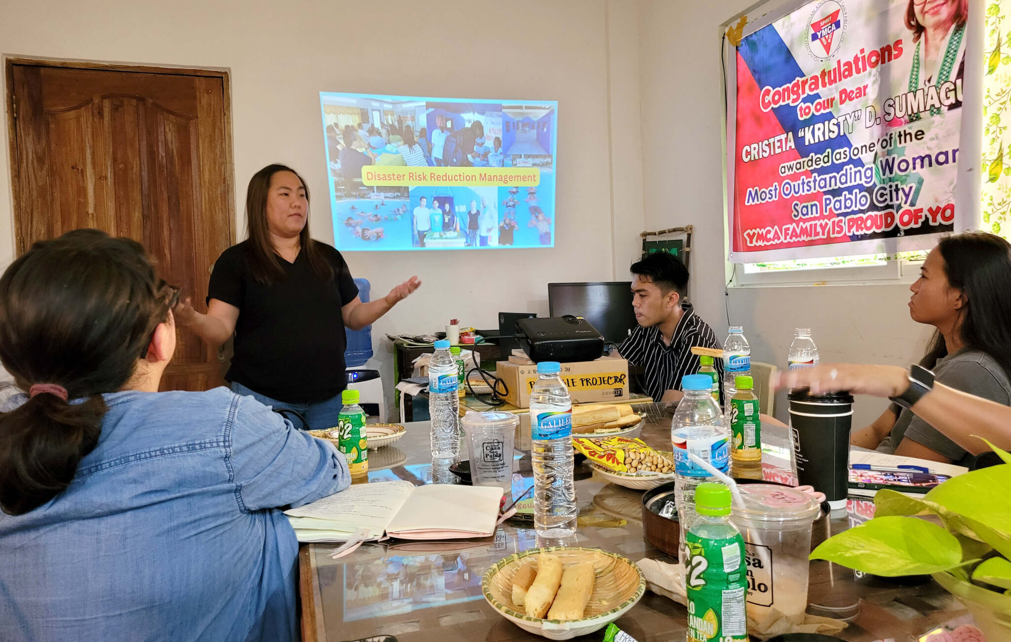 A woman is standing in front of a room presenting information to people sitting down at a table.
