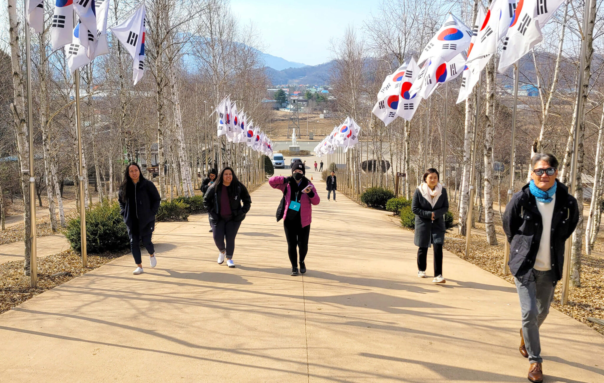 A group of five people are walking uphill on a paved path. Each side of the path is lined with South Korean flags. It is bright and sunny.