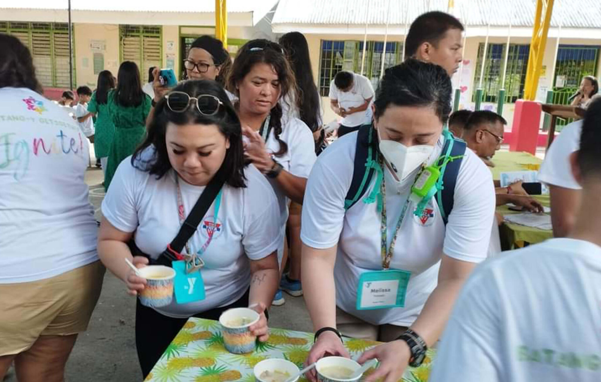 Two women are picking up cups of food from a table. They are serving the food to students at an elementary school in San Pablo City in the Philippines.