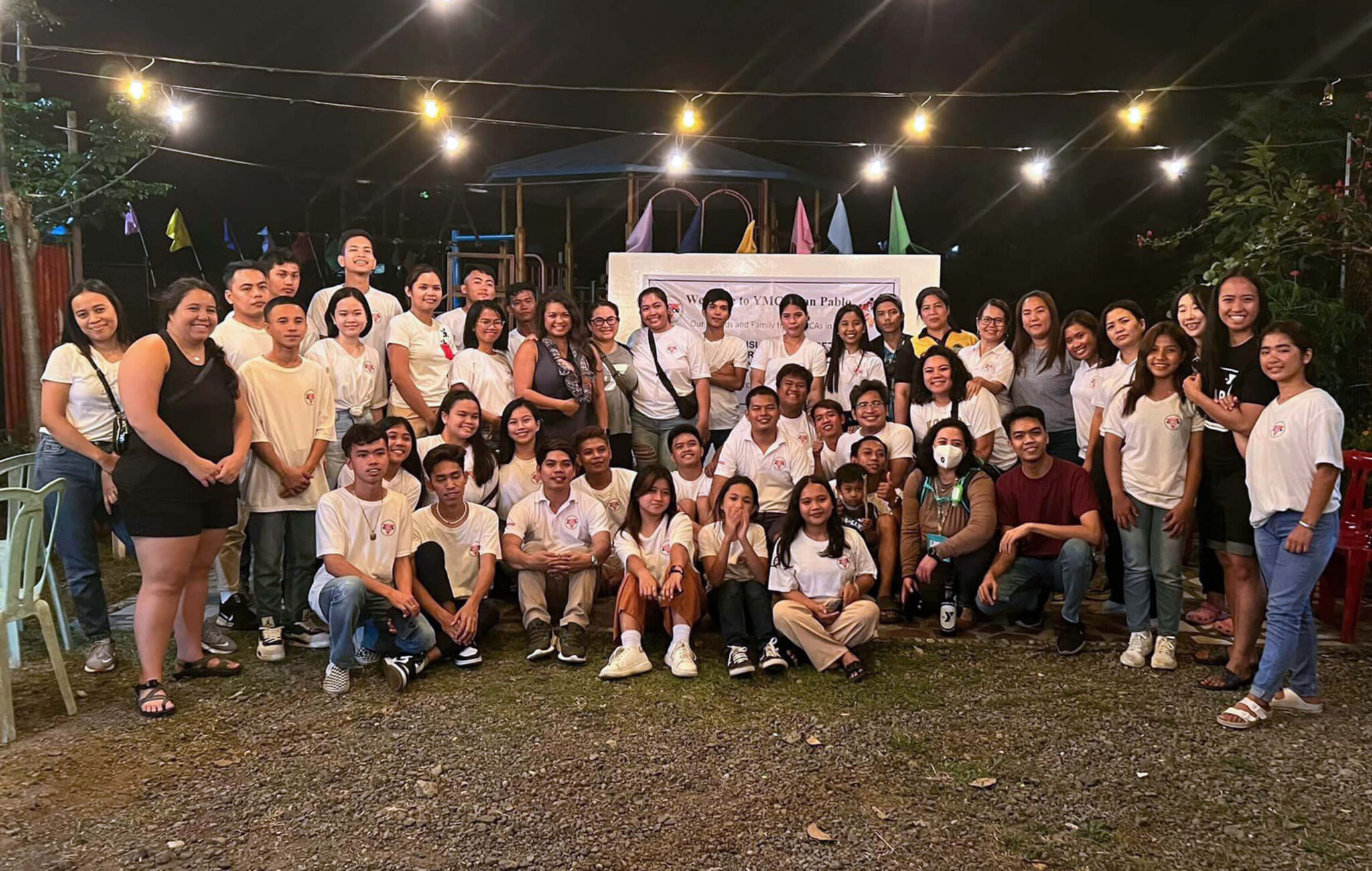 A large group of people gather for a group photo underneath string lights.