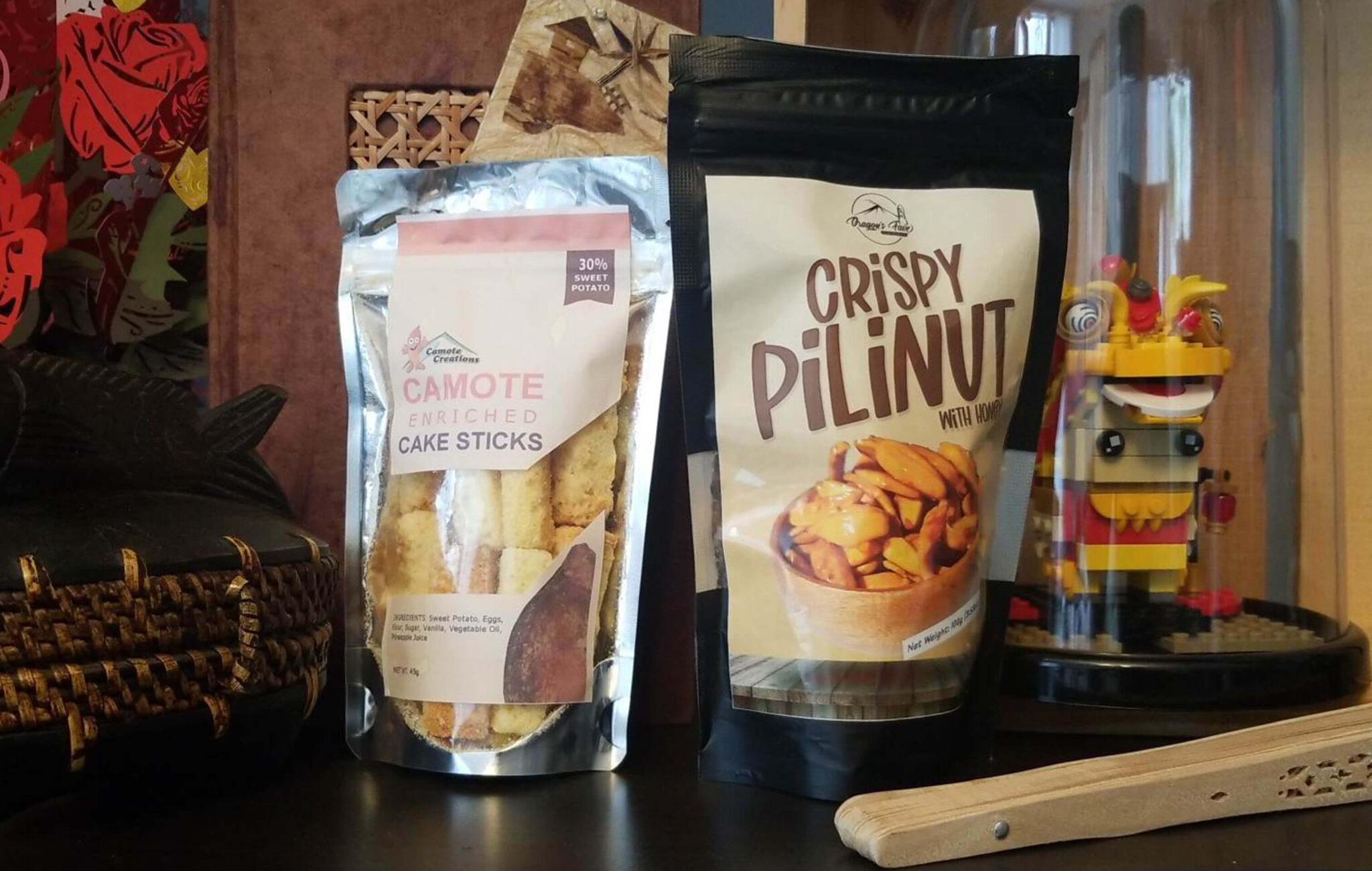 Two packages of food are placed on a shelf. One package is of processed cake sticks and the other is of crispy pili nuts. In the background are various objects from the Philippines.