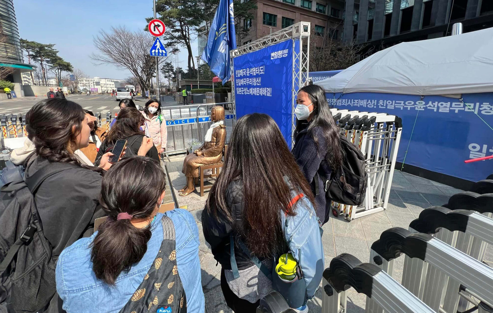A group of women are gathered around a statue that is seated in front of a blue background with Korean text on it.