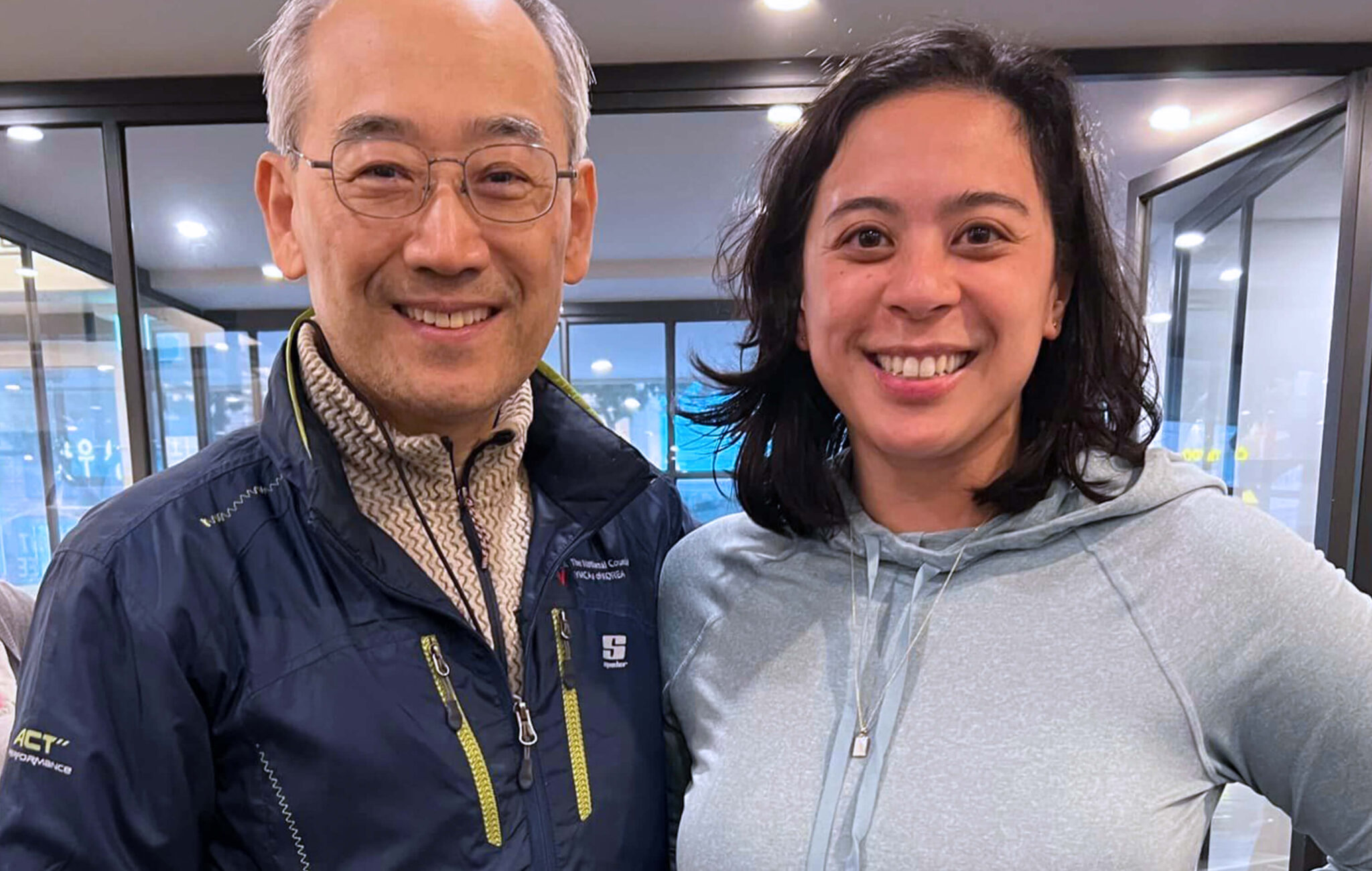 An Asian man wearing glasses and a navy blue zip down jacket poses with an Asian woman with a light blue hoodie. They are both smiling.