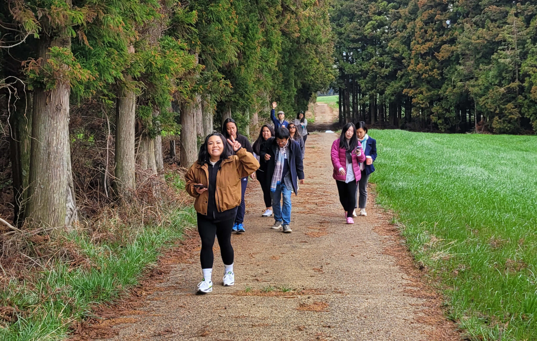 A group of people are walking uphill on a dirt trail next to a tree line. The person at the front of the group is showing a peace sign with her fingers and smiling.