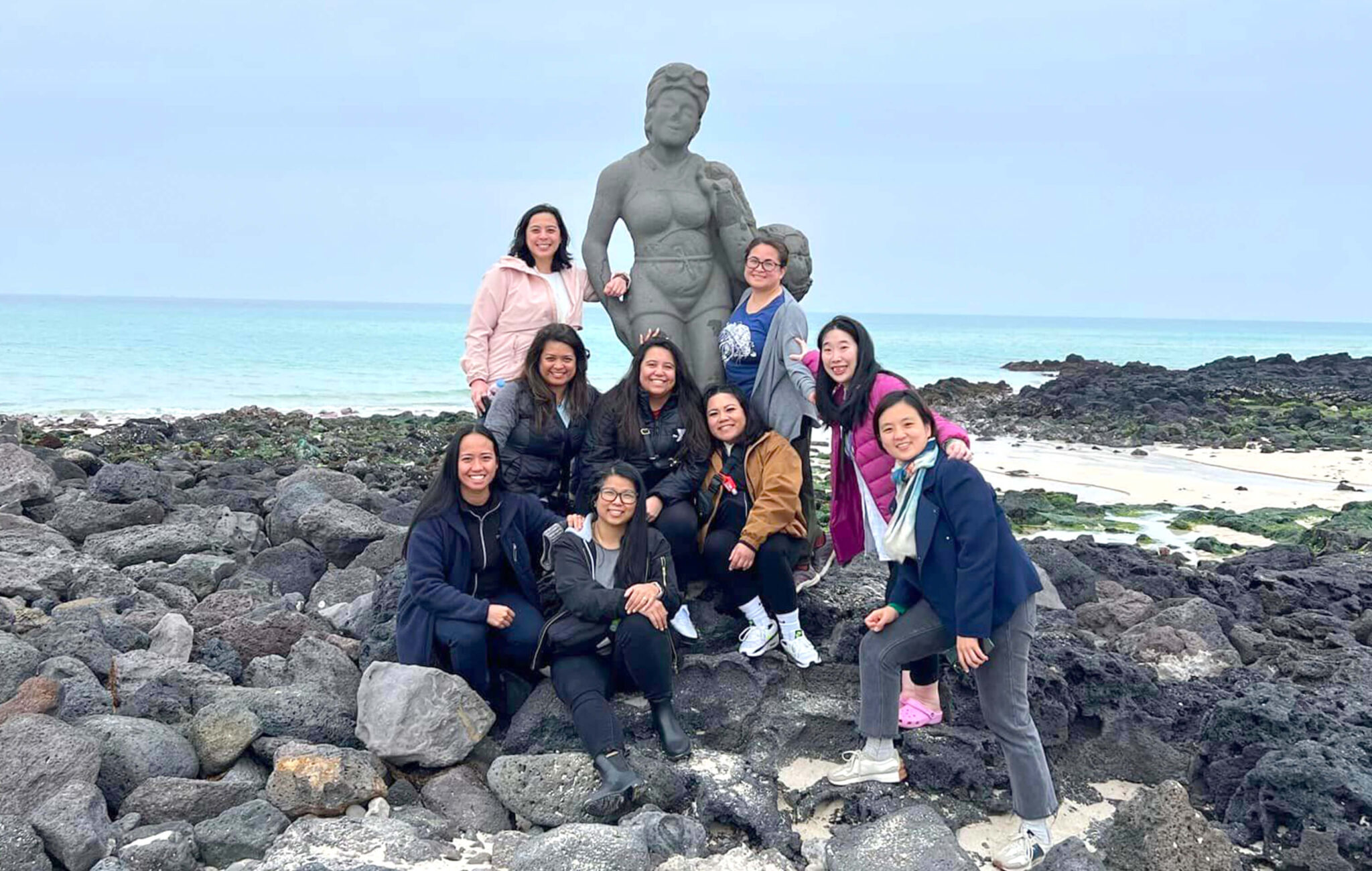 Nine women are standing on rock formations on a beach. They are gathered around a stone statue of woman that represents the hae-neyo women divers on Jeju Island.