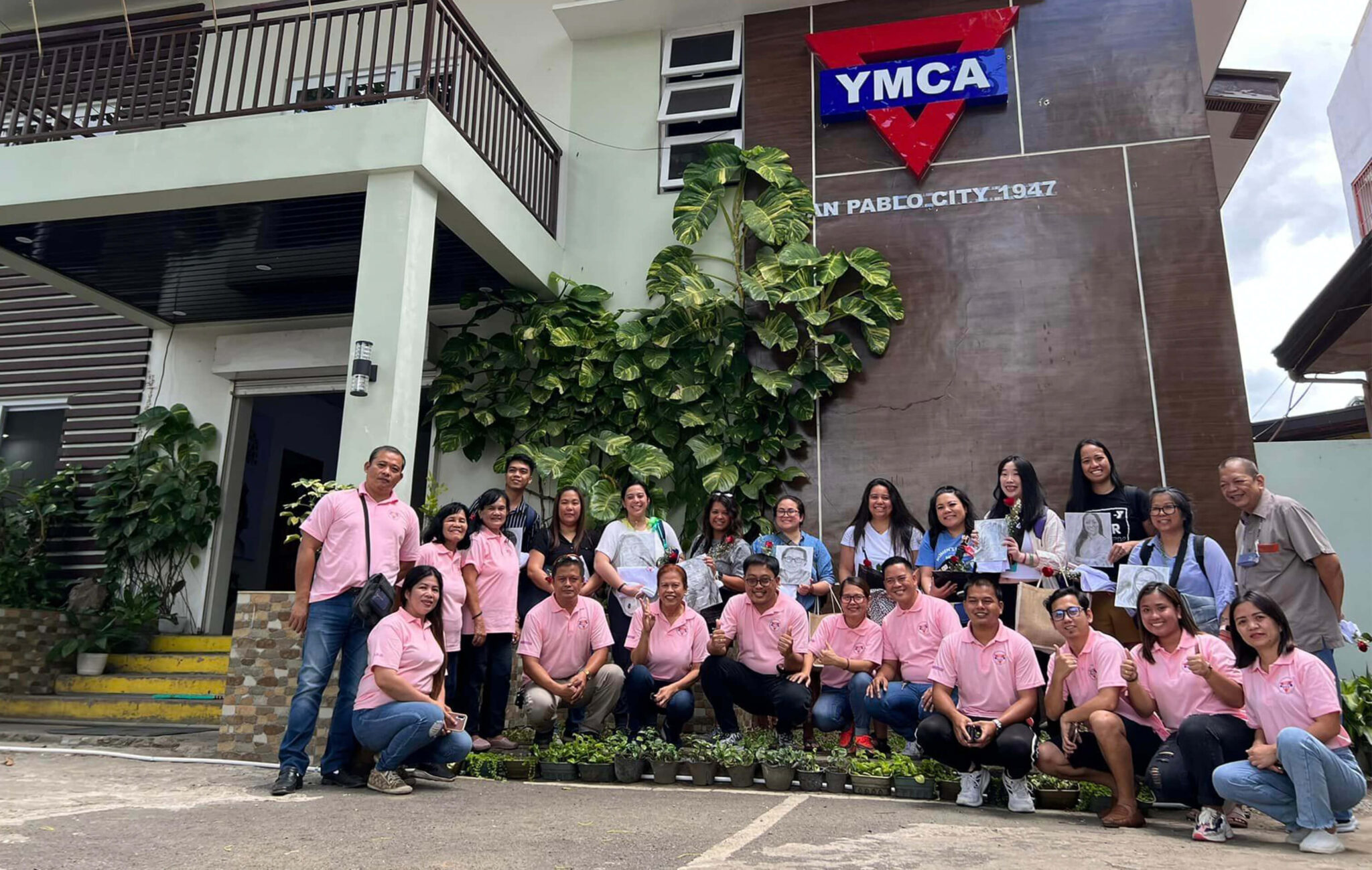 A group of people gather in front of a building. Some are kneeling in a row with pink polo shirts. The people standing behind them are wearing various clothing. The building has a YMCA logo sign on it with another sign that reads, "San Pablo City 1947" below it.
