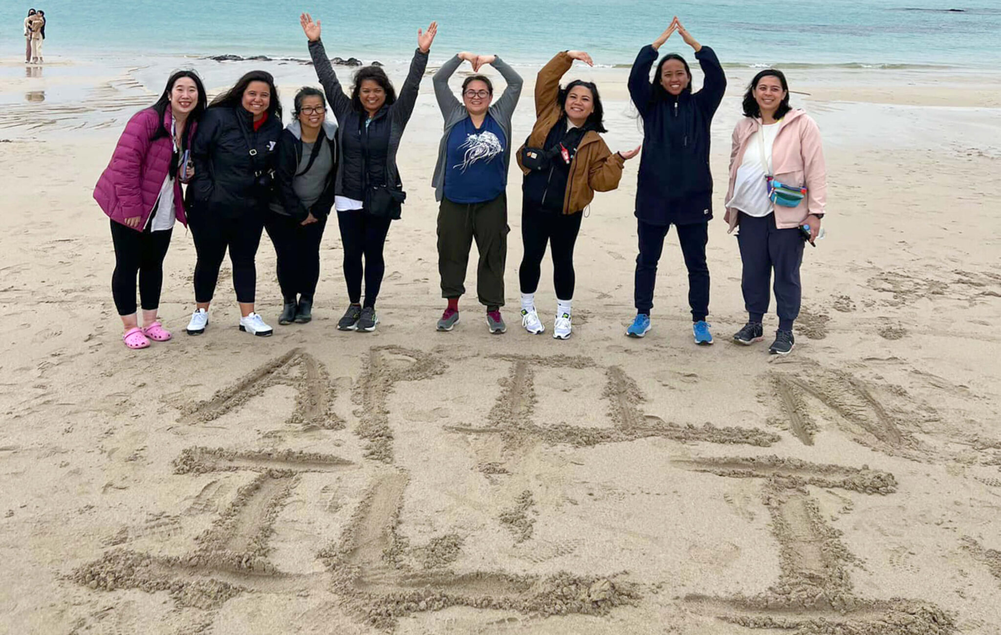 Eight women standing side by side on the sand at a beach on Jeju Island. Four of the people are using their arms to spell out "Y," "M," "C," and "A." Written in the sand is "APILN ILI."