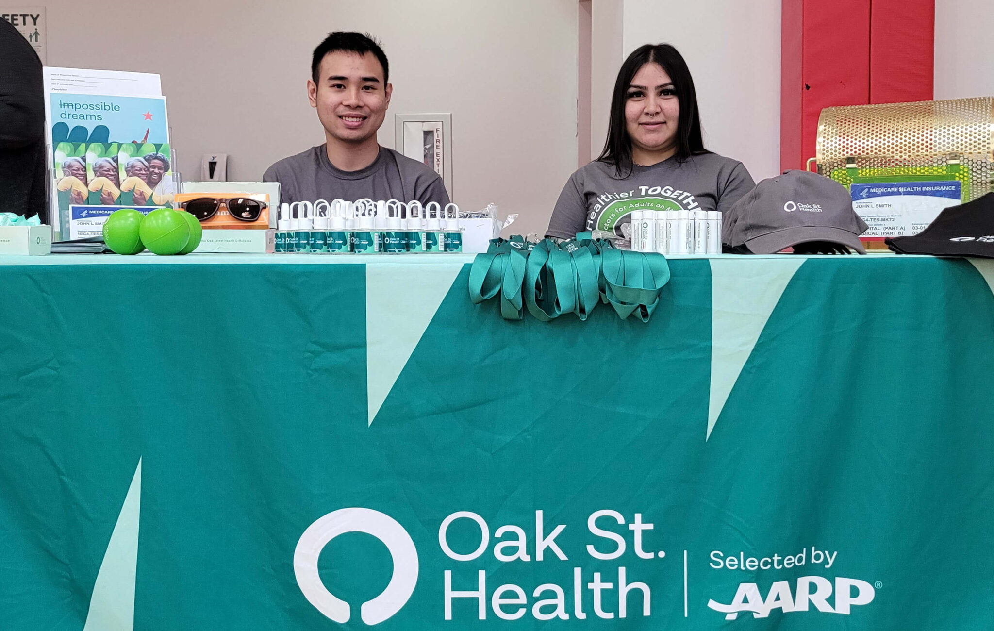 A man and a woman are seated behind a table. The table is covered by tablecloth with "Oak St Health" printed on it. The table has various items available to attendees of Community Day, like hand sanitizer and lip balm.