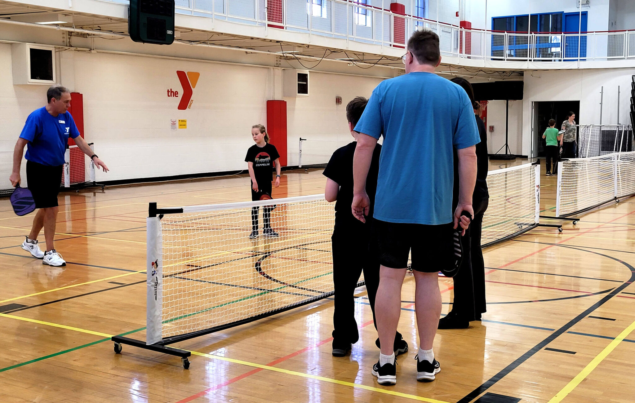 A YMCA staff member is teaching a group of four people about pickleball. The instructor is standing on one side of the net with one participant while the other three are standing on the other side of the net on a pickleball court. The court is in a large gym.