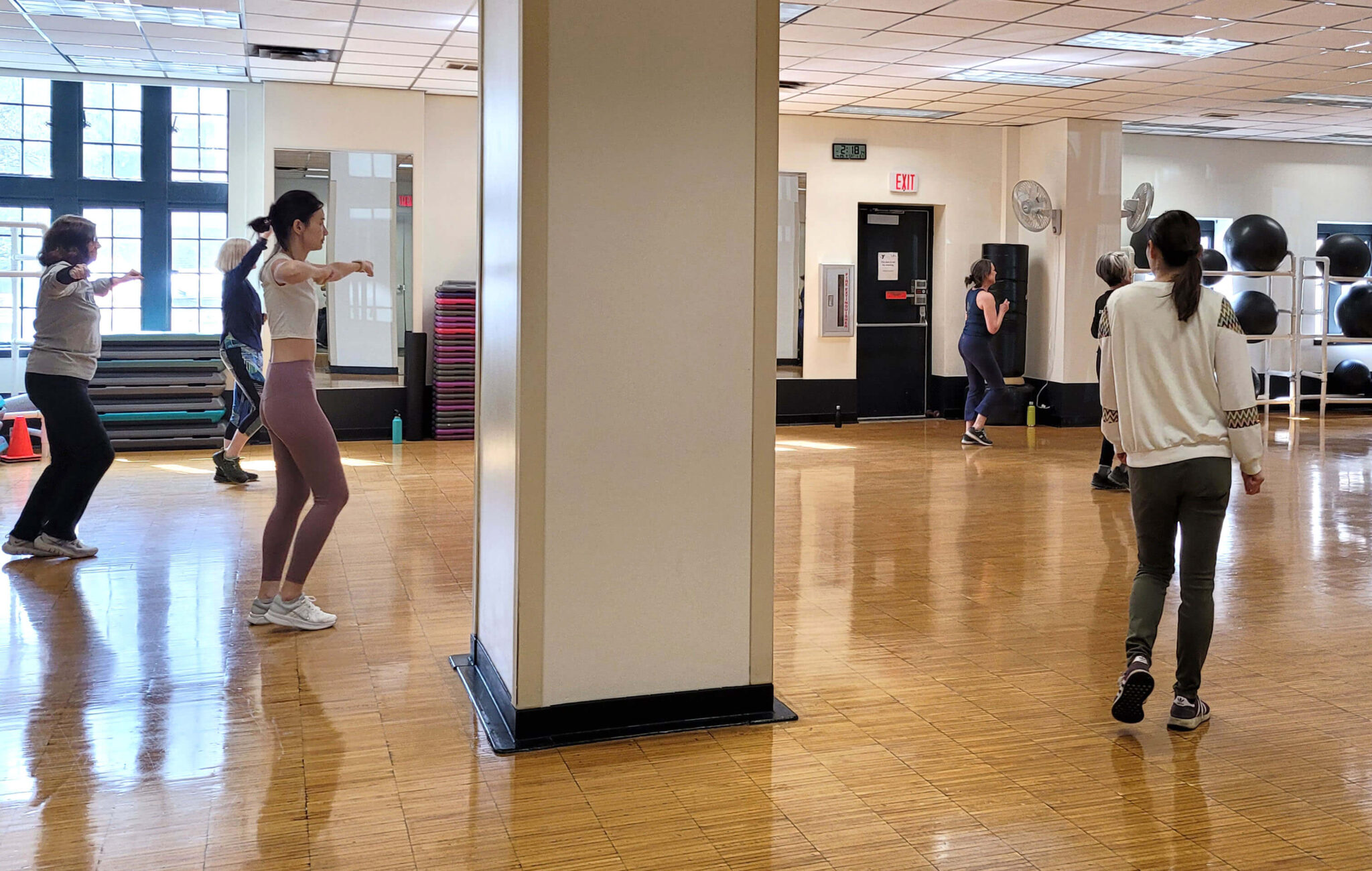 Women are in the Group Fitness studio. Three of the women are in a dance position, following along with the Zumba instructor (not pictured). Another woman is walking towards the group to join the class. There is a large structural column in the room.