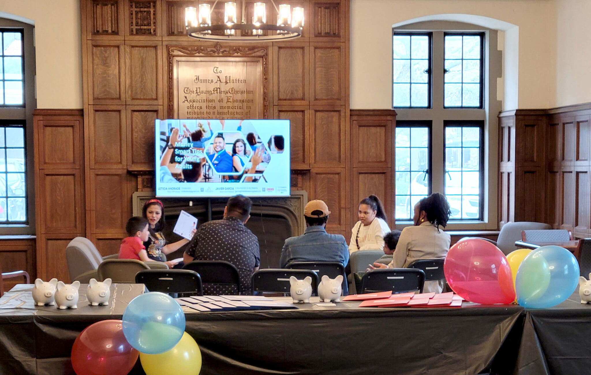 A group of seven people are seated in a circle. Some of the people are children and others are adults. There is a monitor displaying the words, "Money Smart Tips for Young Adults" in the room. The room also has tables with black tablecloths that have folders of printed materials laid out on them. There are also piggy banks and balloons on the tables. The room has wood paneling along the walls.