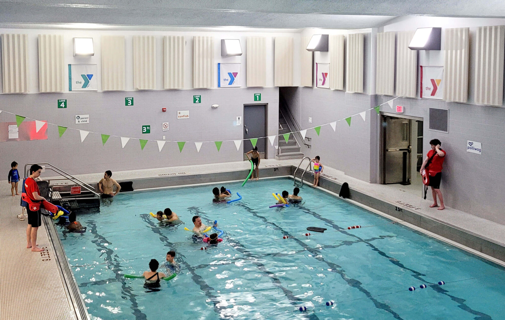 Multiple groups of families are in a swimming pool. Some have pool noodles and other tools they're using. There are two lifeguards on each side of the pool carrying equipment.