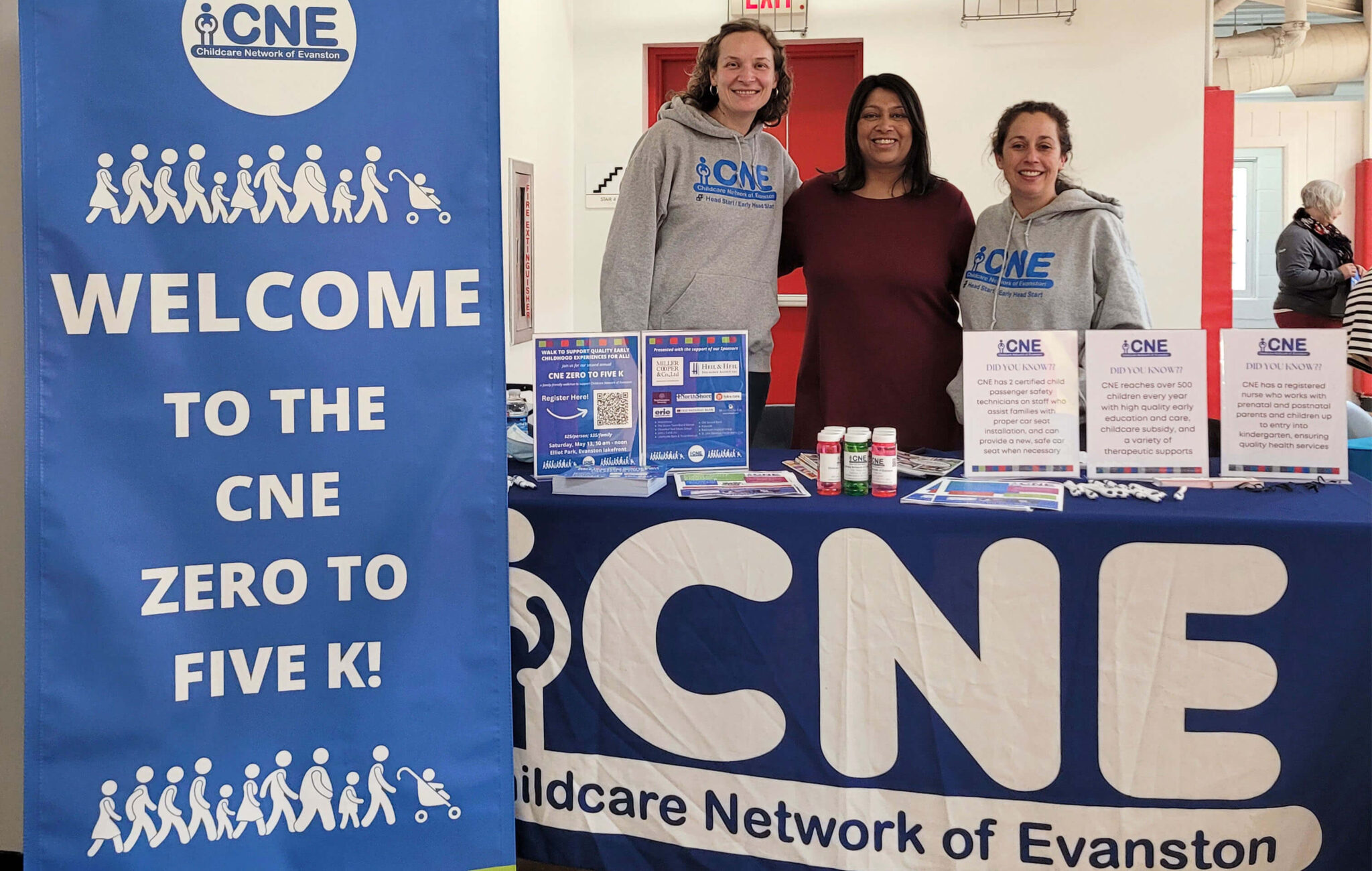 Three women are standing behind a table. There is a pop-up banner next to the table that reads, " Welcome to the CNE Zero to Five K!" The tablecloth has a large logo for the Childcare Network of Evanston. The table has printed resources available to attendees like flyers and informative data points about child safety an education.