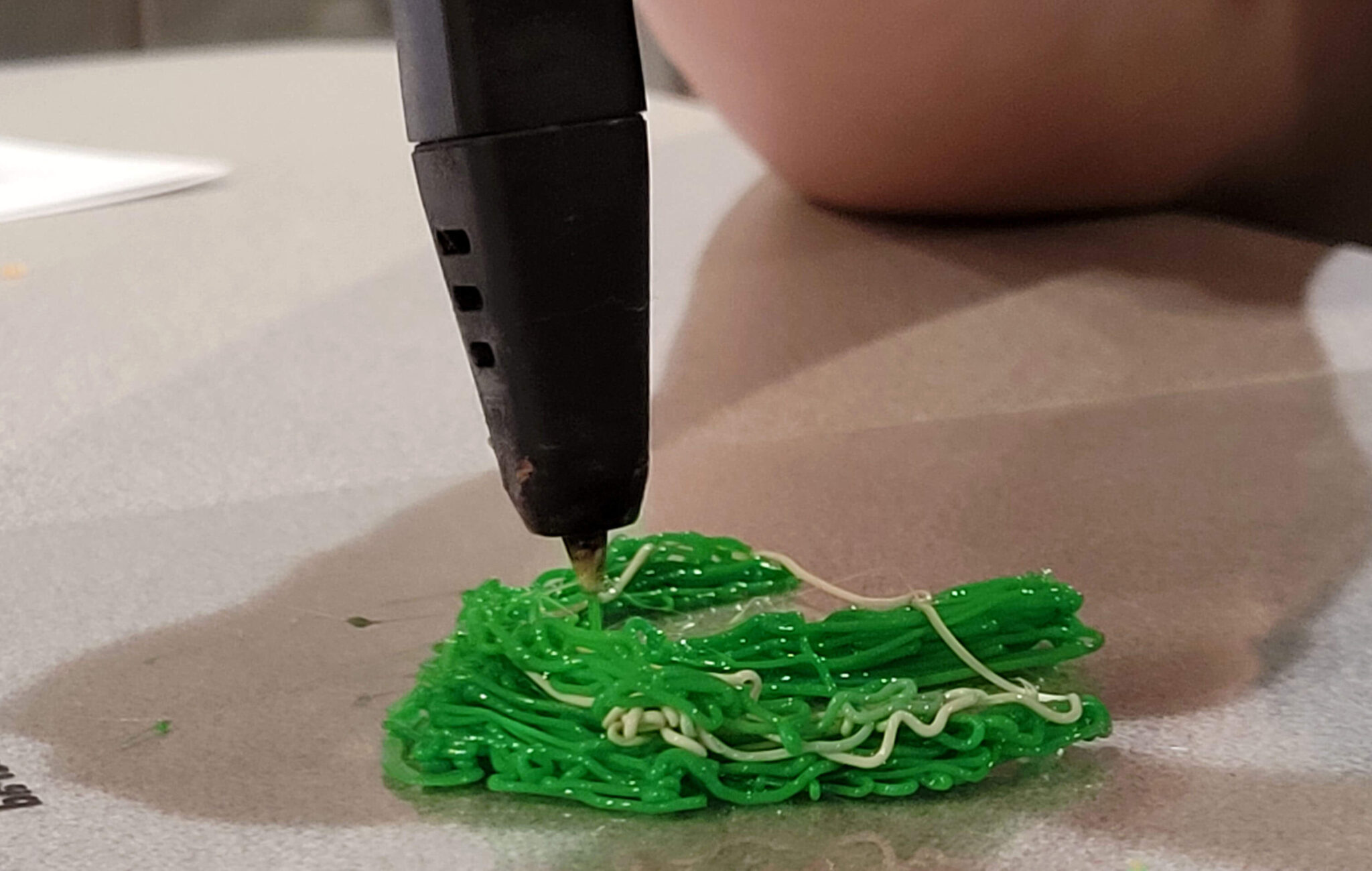 This is a very close up picture of a 3D printing pen. The pen is printing a green plastic thread that is a blob-like structure.