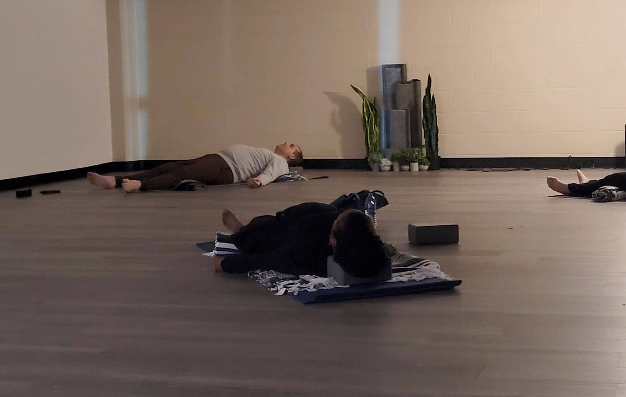People are laying on their backs on top of yoga mats and blankets for support. There are additional yoga props nearby to each person. The room is dimly lit.