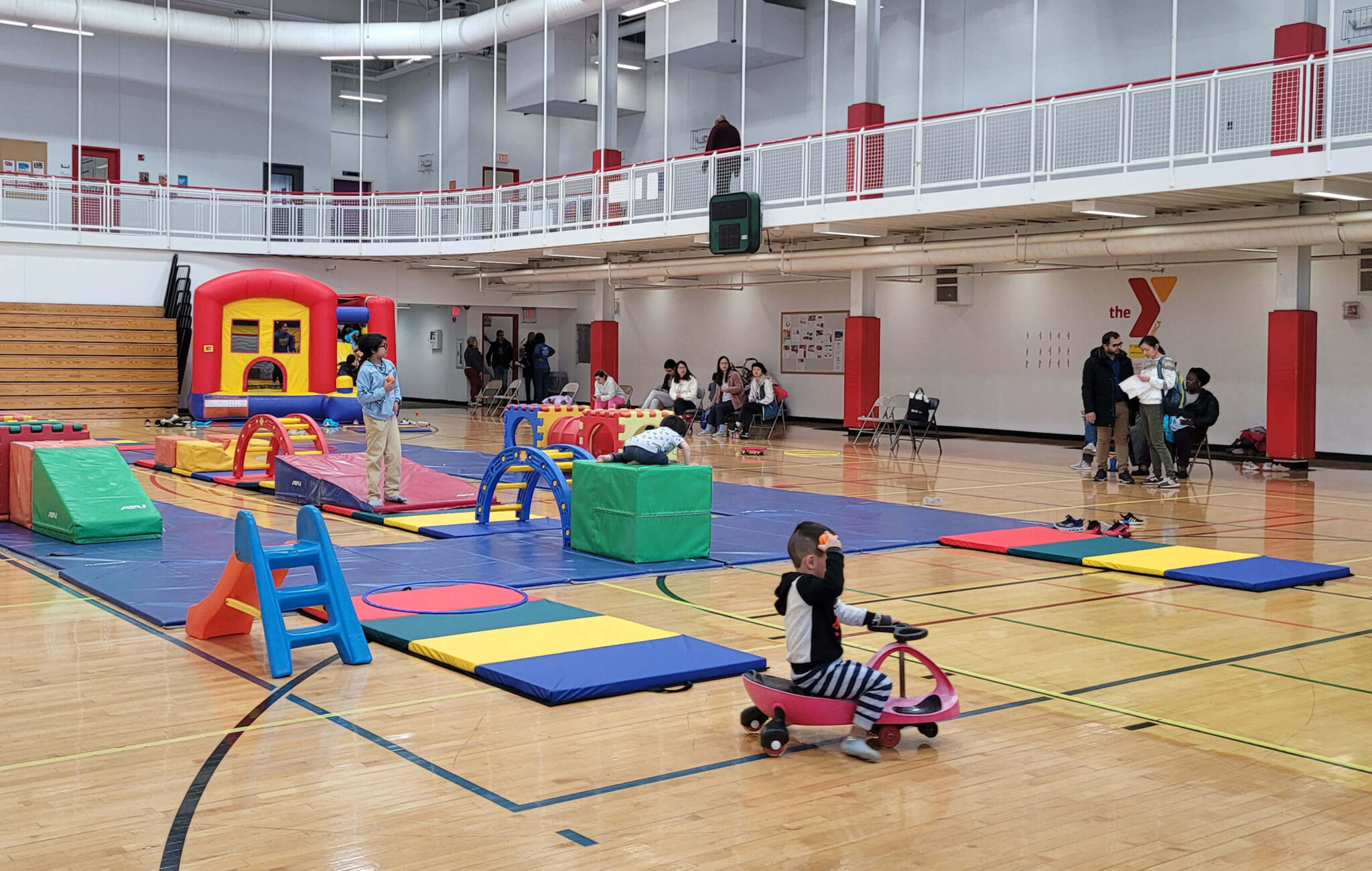 Brightly colored gym mats are laid out on a gym floor with modular jungle gym pieces are strategically placed throughout the space. Parents are seated in folding chairs along the perimeter of the gym watching their kids play.
