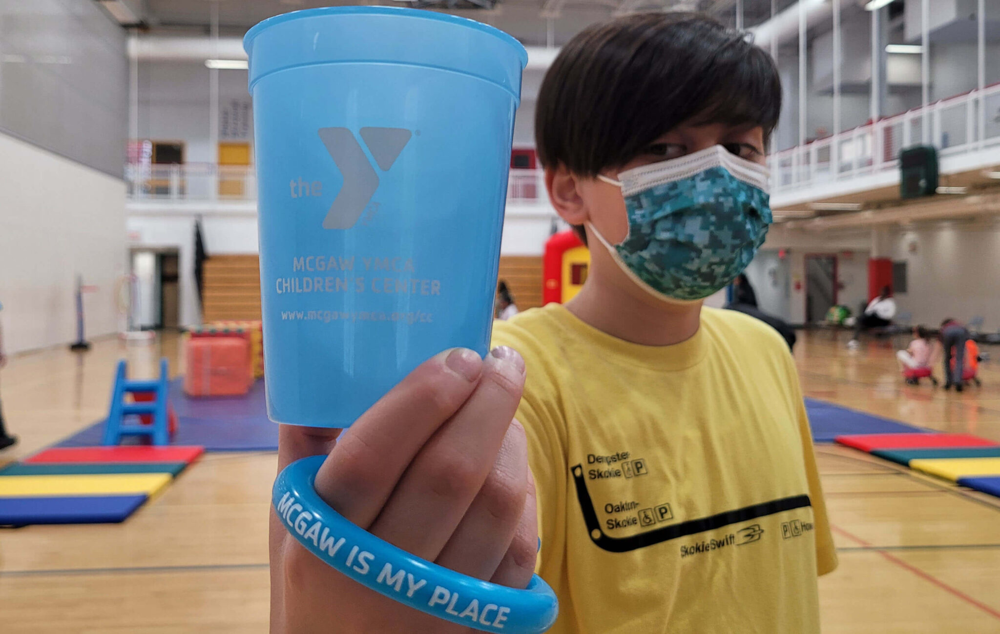 A young man is wearing a yellow t-shirt and holding a blue cup in his hand. The cup has the YMCA logo on it. The young man is also wearing a blue bracelet that reads, "McGaw is my place." He is standing in a gym in front of a play area set up with gym mats.