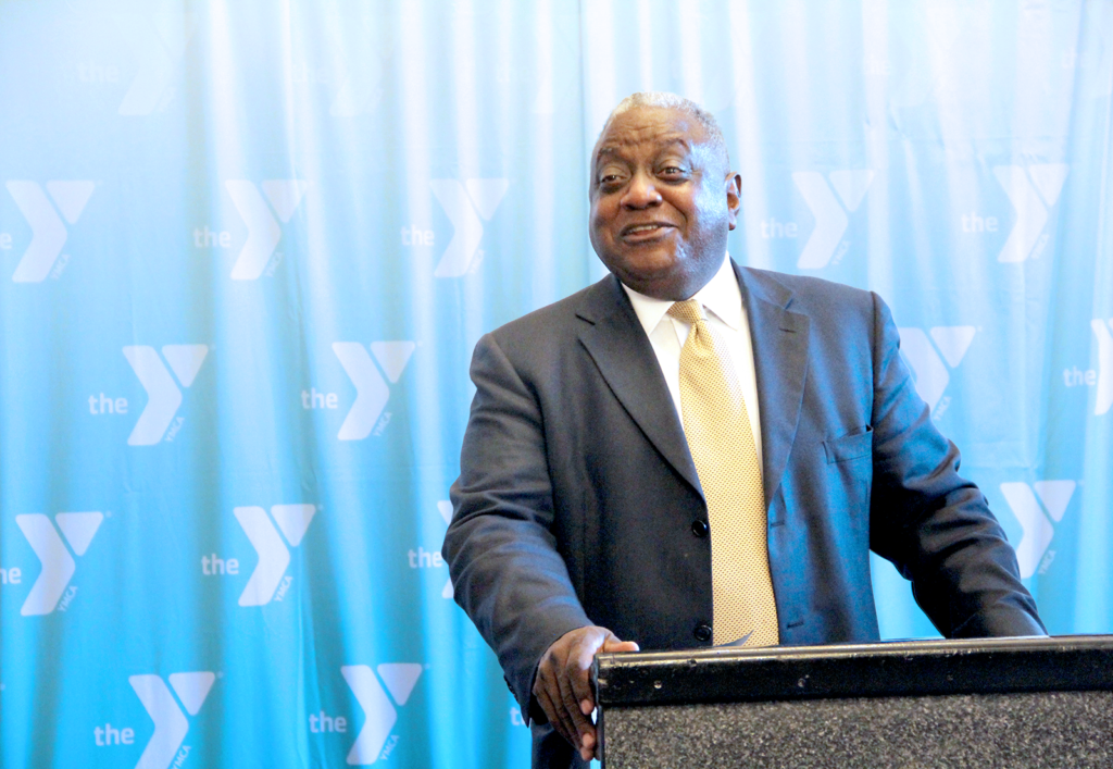 Mark Dennis speaking at a podium, looking off to the side smiling, with YMCA logos in the background.