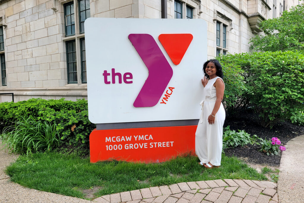 Tai is standing in front of the YMCA sign outside of the main building at the corner of Maple and Grove. She is wearing a white pantsuit and is smiling. 