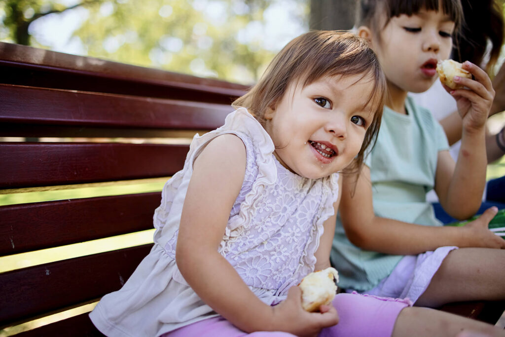 A young child is sitting on a bench with a sandwich in their hand and they are smiling. They are sitting next to a child who is eating a sandwich. 
