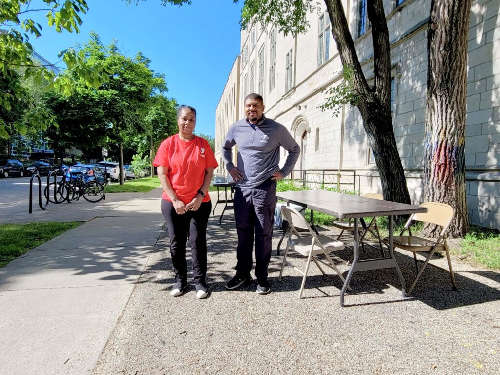 McGaw YMCA staff offer free summer meal pickup outside of the main building at the corner of Grove Avenue and Maple Street. They are standing next to a table with chairs and smiling. The person on the left is wearing a red t-shirt and black pants and the person on the right is wearing a grey half-zip t-shirt with black pants. 