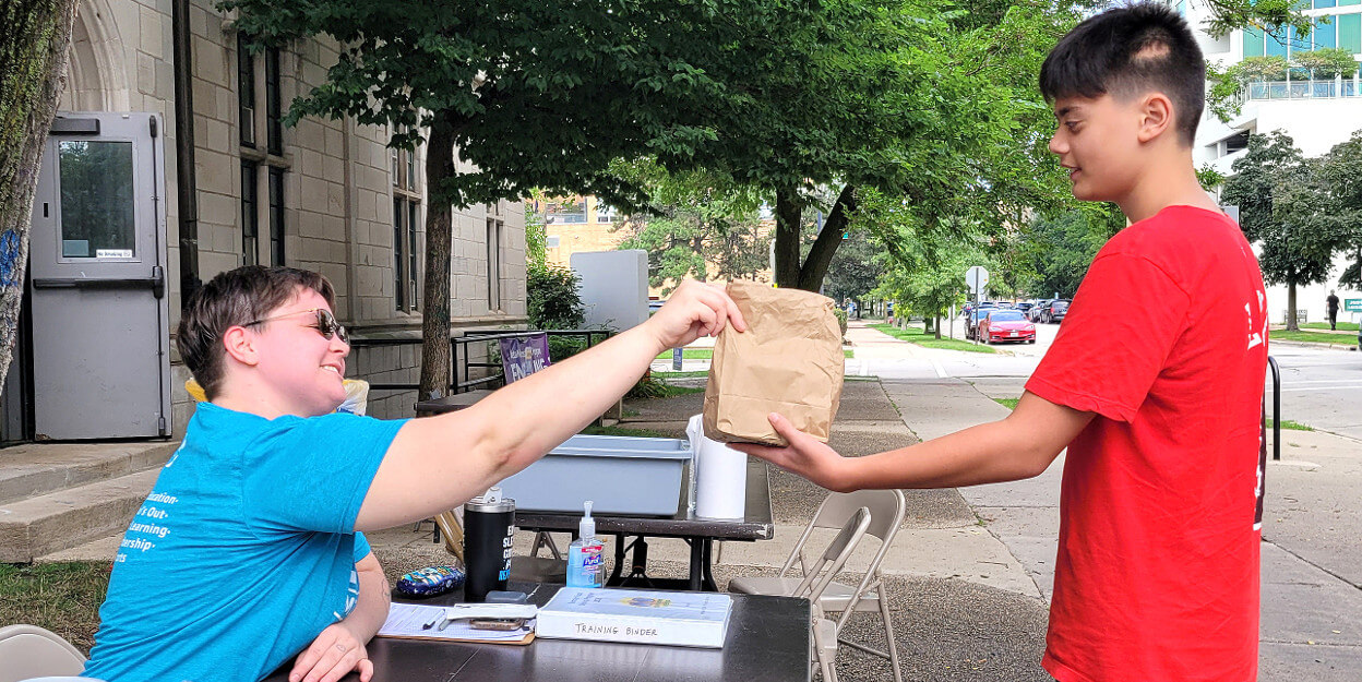 A YMCA staff member is smiling and sitting at a table, handing a bag lunch to young adult. They are outside. 