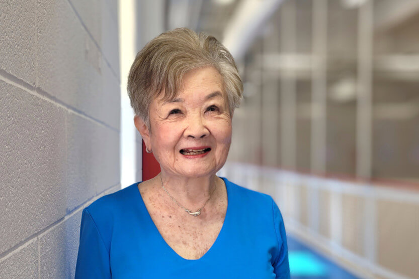JoAnn is standing on the indoor track, leaning against a white brick wall, and wearing a blue v-neck top.