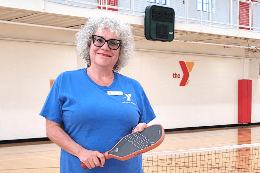 Liette is wearing a blue short sleeved Y staff shirt while holding a pickleball paddle in her hand.
