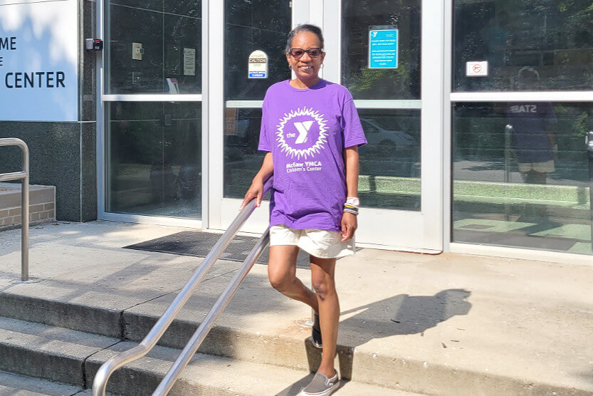 Chantel is wearing a purple McGaw YMCA Children's Center t-shirt and light colored shorts. She is standing on the front staircase of the Children's Center, with her hands on the railing while smiling.