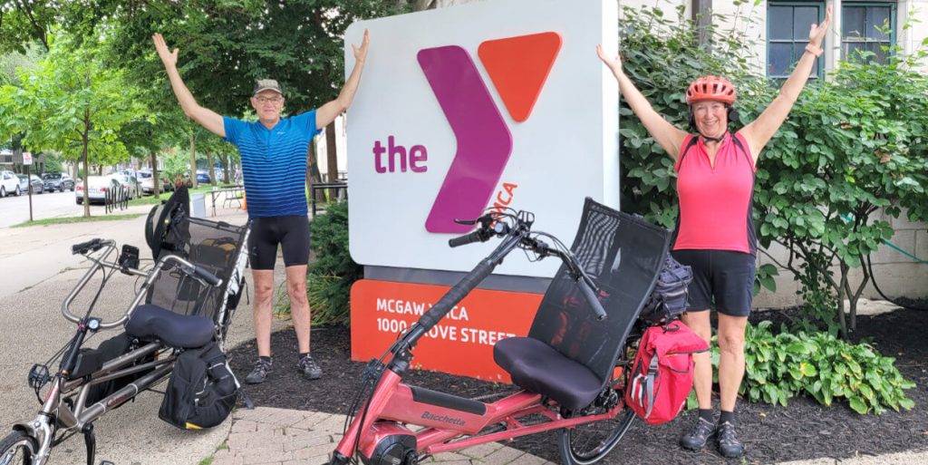 Mark and Noreen posing in front of the YMCA logo in front of the Main Building. Their bikes are in front of them and their arms are up in a Y shape.