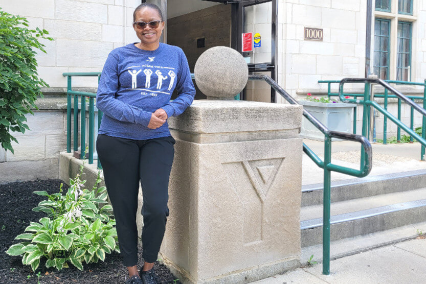 Brenda is standing in front of the McGaw YMCA with sunglasses, black pants, and a long-sleeve YMCA t-shirt on.