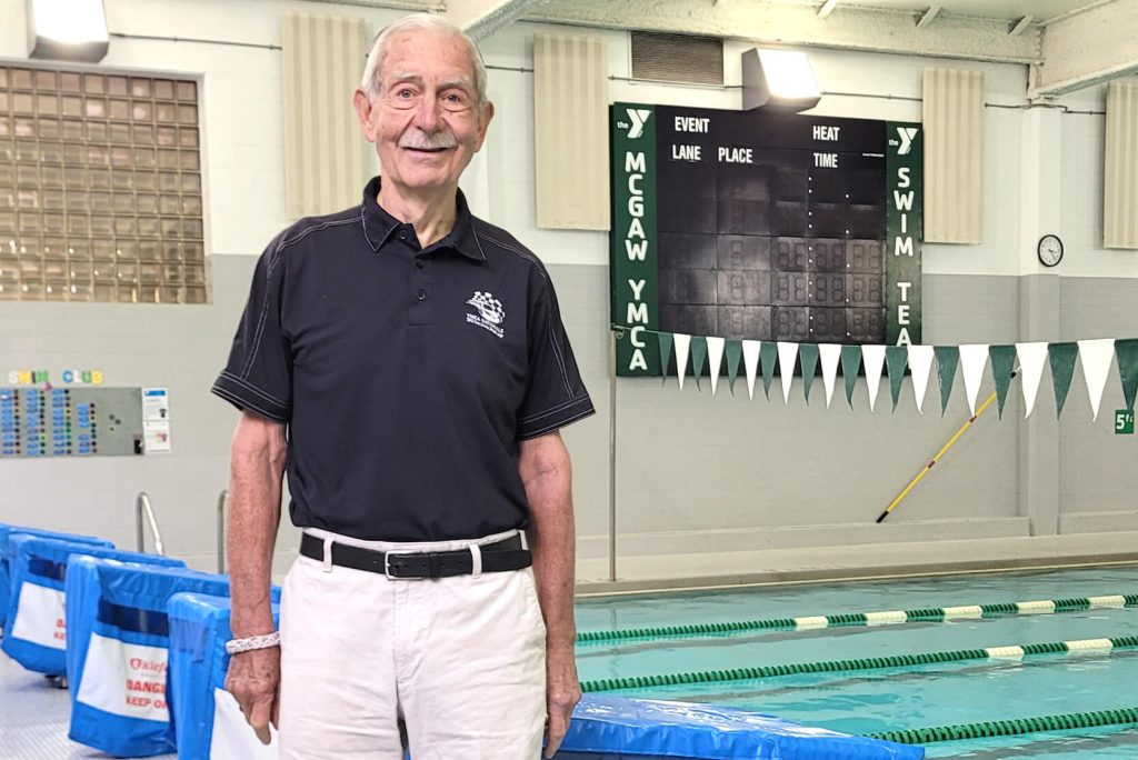 Richard is standing on the pool deck and smiling. He's wearing a black polo and khaki shorts with the MYST scoreboard in the background.