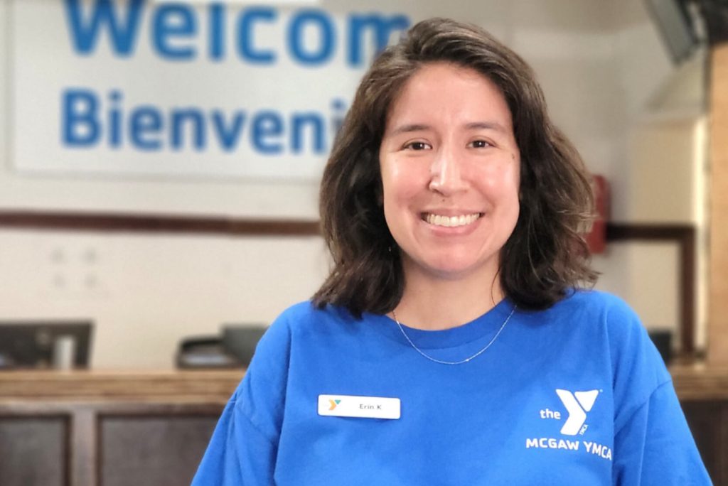 Erin is wearing a blue McGaw YMCA t-shirt and smiling. She is standing in the Main Lobby, in front of the front desk.