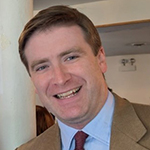 Christian has short brown hair. He is smiling and wearing a tan-brown blazer with a light blue button shirt and a tie.