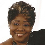 Deldri is smiling and wearing dangling earrings, a necklace, and a black top. The background is white.