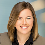 Erinn is wearing a black button down under a grey-brown blazer. She has brown hair and it sits at her shoulders. The background is a pale blue with streaks of a cream color.