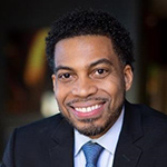 Henry is wearing a black blazer with a white button down shirt and a blue tie. He has short black hair and has some facial hair. The background is very dark.
