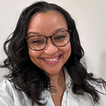 Krenice is smiling and wearing eye glasses. Her long black hair is down and she is wearing a white button down shirt. She is smiling.