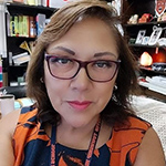 Mercedes is wearing eye glasses and wearing a navy and orange floral top. She is in an office and in the background are books and shelves.