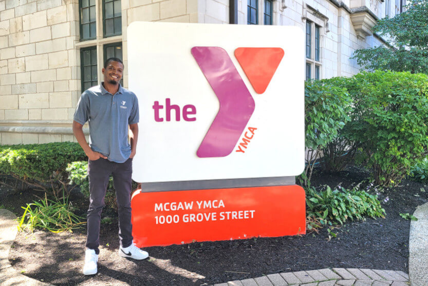 Eric is standing next to a Y sign that is outside of the Main Building. He is smiling and wearing a blue-grey staff polo and black pants.