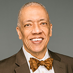 Theodore is wearing a grey-brown blazer, white button shirt, and a bow tie. He is smiling and the background is light grey.