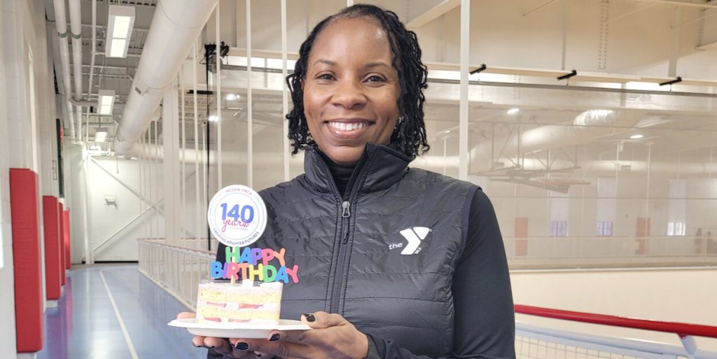 President/CEO Monique Parsons is standing on the upper level track of the Sebring-Lewis Center holding a piece of cake with "Happy Birthday" and "Celebrating 140 years in Evanston" decoration. She is wearing a black shirt and with a black puffy vest with a YMCA logo on the chest. She is smiling.