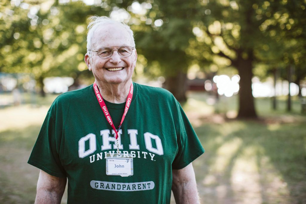John is standing on the Camp Echo property, smiling, and wearing a green Ohio University Grandparent t-shirt