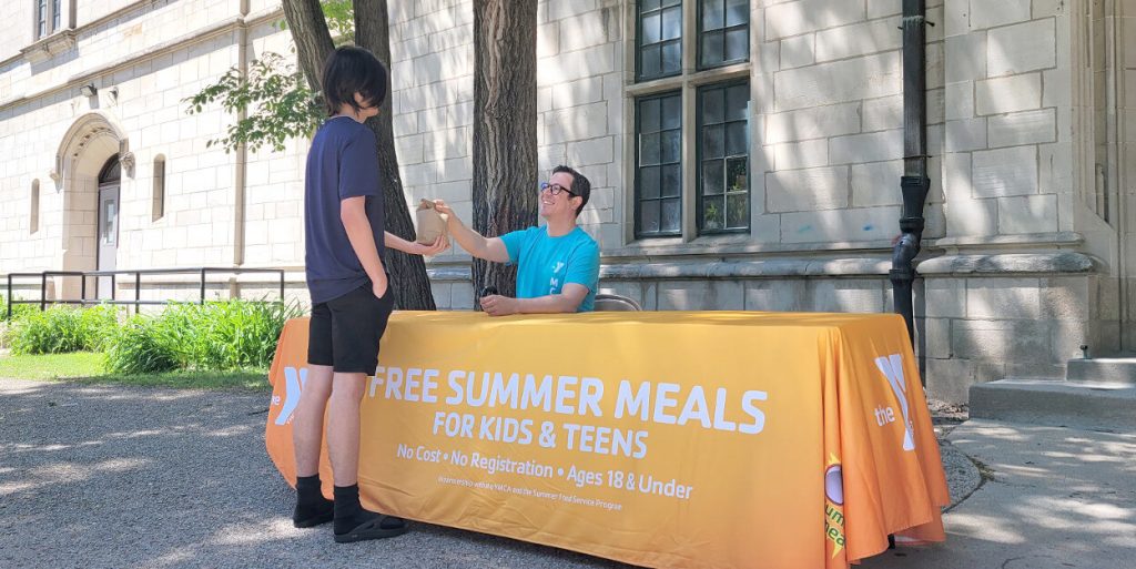 A YMCA staff person is seated at a table while handing a sack lunch to a teenager.