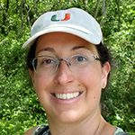 Cristi 150 x 150 Cristi is wearing a white University of Miami baseball cap and eye glasses. She is standing in front of greenery and smilling.