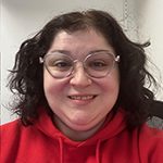 Melanie 150 x 150 Melanie has wavy brown hair and is wearing eyeglasses. She's also wearing a red hoodie and smiling.
