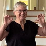 Nancy 150 x 150 Nancy is standing in front of a staircase wearing a black top. She has short grey, white hair and is wearing eye glasses.